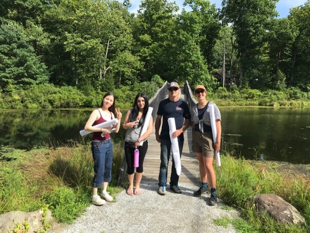 Four Rutgers students with rolls of paper in front of foot bridge with wooded area in background.