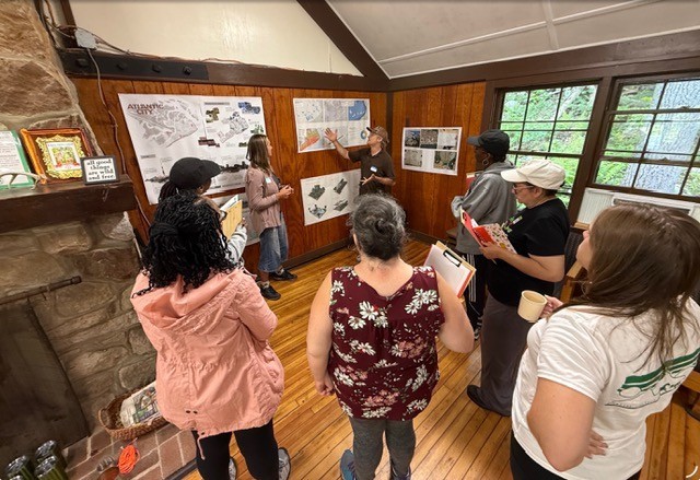 People watching person presenting posters in wood floored and paneled room near a fireplace.