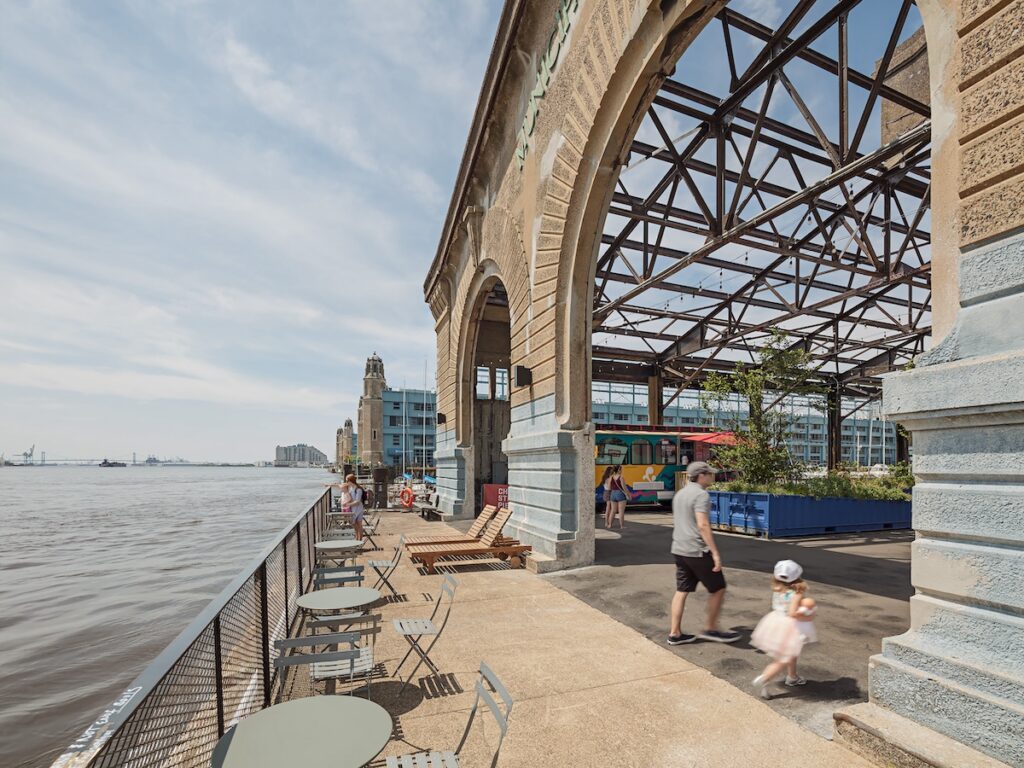 Photo of the waterside end of Cherry St Pier with Delaware River to left, walkway with seating in the middle, and to the right are arched openings leading into sitting area with food truck and plantings in shipping containers. The roof of the sitting area is open to sky but with historic industrial trusses intact.