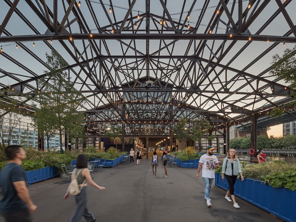 Sitting area of the water-end of the Cherry Street Pier. Two sides and roof are open with only structural elements on sides and metal roof trusses above. Numerous blue shipping containers hold plantings. To the far end is the entrance to the covered area that leads to the street.