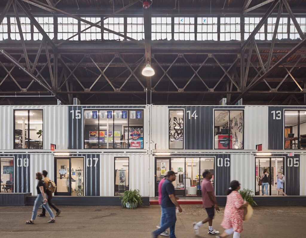 Covered area of Cherry Street Pier containing, along one side, two levels of shipping container art studios, each with large glassed doors and windows. People walk in the foreground.