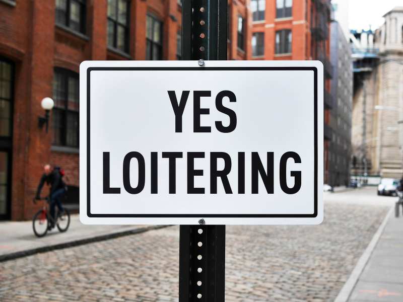 Photo of "Yes Loitering" street sign in foreground with urban street scene behind.