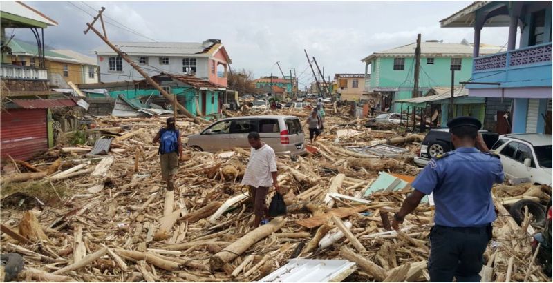 Street covered in wood debris and other parts of structures, leaning utility pole, a few brightly colored standing houses and people walking amongst it.