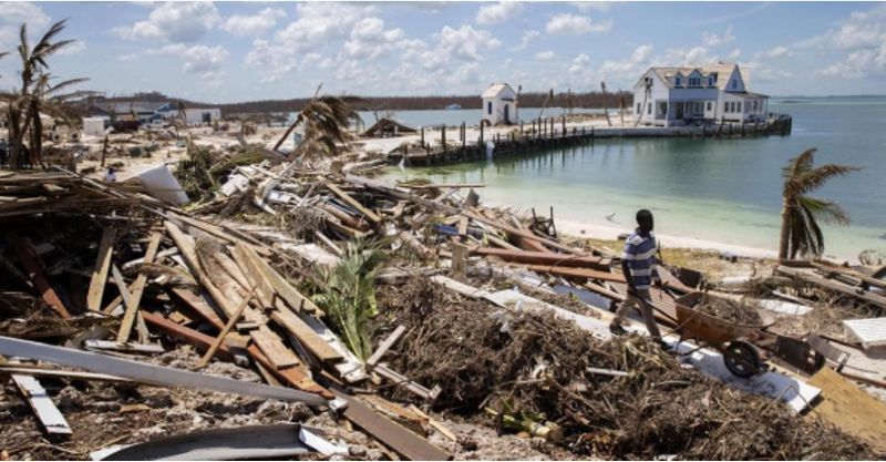 Beach lined with boards and other debris. Man carting debris. Pier in background surrounded by blue-green water.