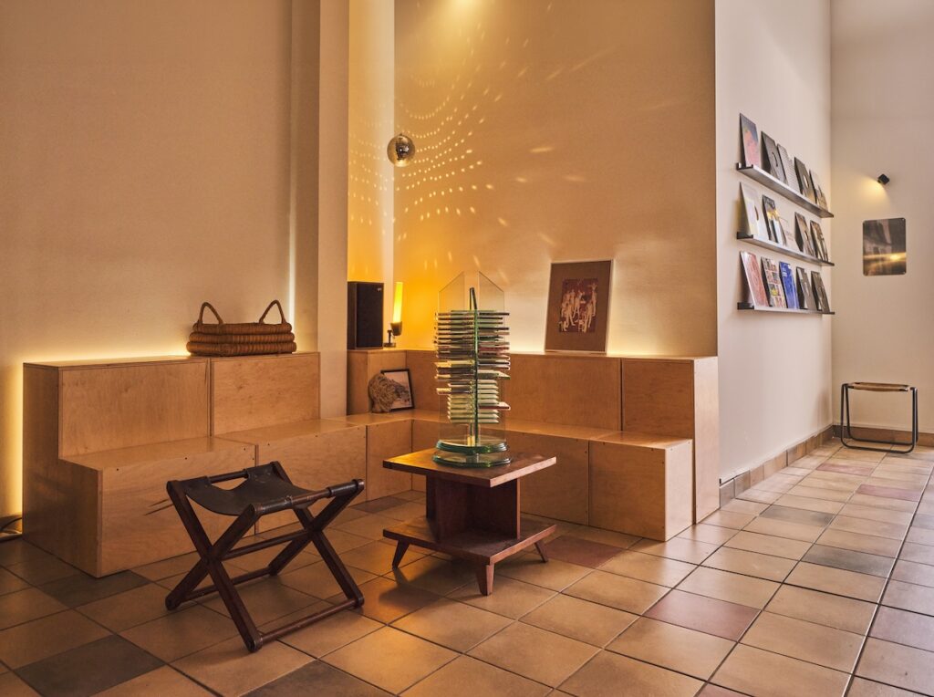 Sitting corner with plywood bench seating in background, wood tables in front, and to side are shelves lined with record albums showing covers. The room is lit with amber lighting from behind the benches and a candle type lamp casting yellow speckles on the wall behind. A handled basket, speaker, lamp and art top the benches.