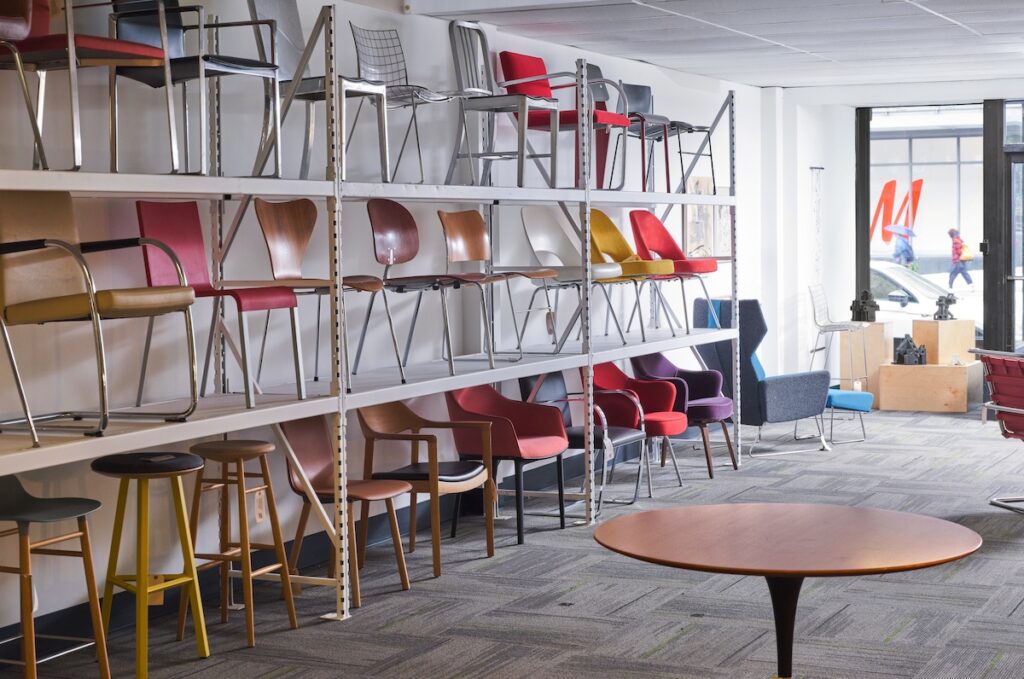 Bright area with glass front and along left wall, a shelving unit with three levels of rows of various stylishly crafted and multicolored chairs and stools.