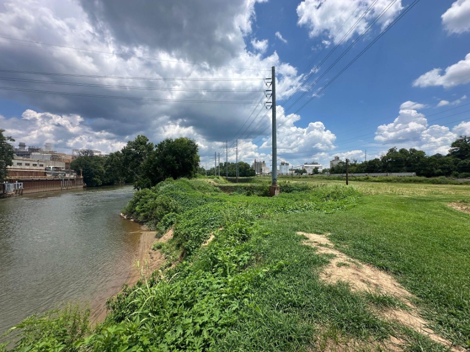 Waterway with industrial building to left, vegetation covered water edge to right with utility poles overhead running into the background.