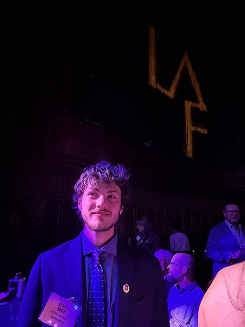 Josh in suit in front of people, LAF logo on dark wall in background.