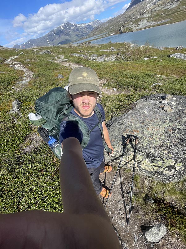 Josh taking long arm selfie with rocky slope to valley with snow capped mountain in background.